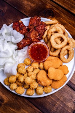 Beyaz tabakta bira için aperatifler servis edilmektedir: tavuk, tempura, soğan halkaları, tavuklu patlamış mısır ve biber sosu