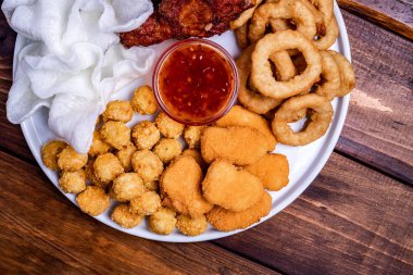 Beyaz tabakta bira için aperatifler servis edilmektedir: tavuk, tempura, soğan halkaları, tavuklu patlamış mısır ve biber sosu