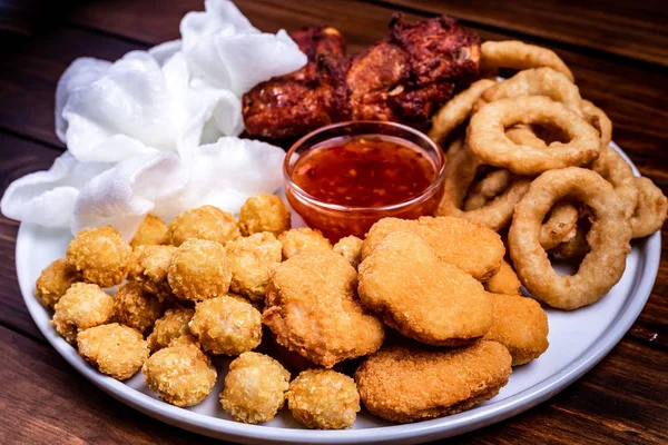 Snacks for beer on a white plate are: chicken nuggets, onion rings in tempura, chicken popcorn and chili sauce