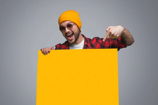 英俊明亮的时髦男子在黄色的帽子和太阳镜指向黄色的海报片上灰色 — 图库照片