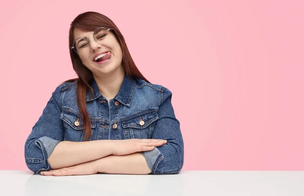 Charming plump girl showing tongue sitting at desk — Stock Photo, Image