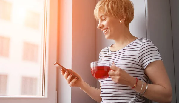 Mujer alegre con té usando teléfono inteligente cerca de la ventana —  Fotos de Stock