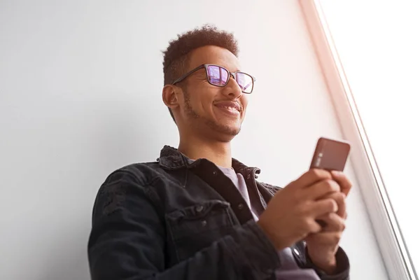 Sorrindo macho preto usando smartphone perto da janela — Fotografia de Stock