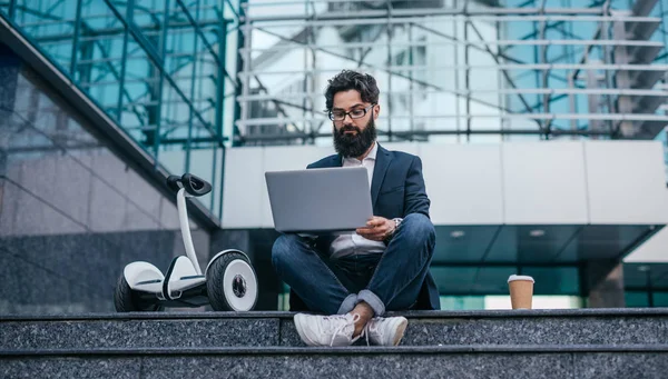 Brodaty Hipster przy użyciu laptopa w pobliżu transportu elektrycznego na zewnątrz budynku — Zdjęcie stockowe