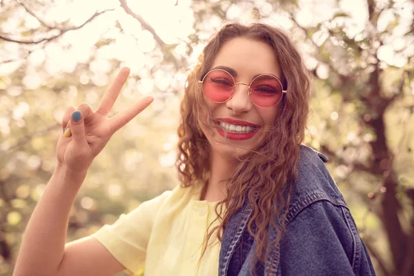 Cheerful trendy woman showing peace sign while standing at park Stock Picture