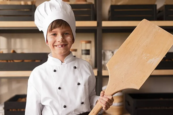 Alegre niño en uniforme de cocinero con pala en panadería —  Fotos de Stock