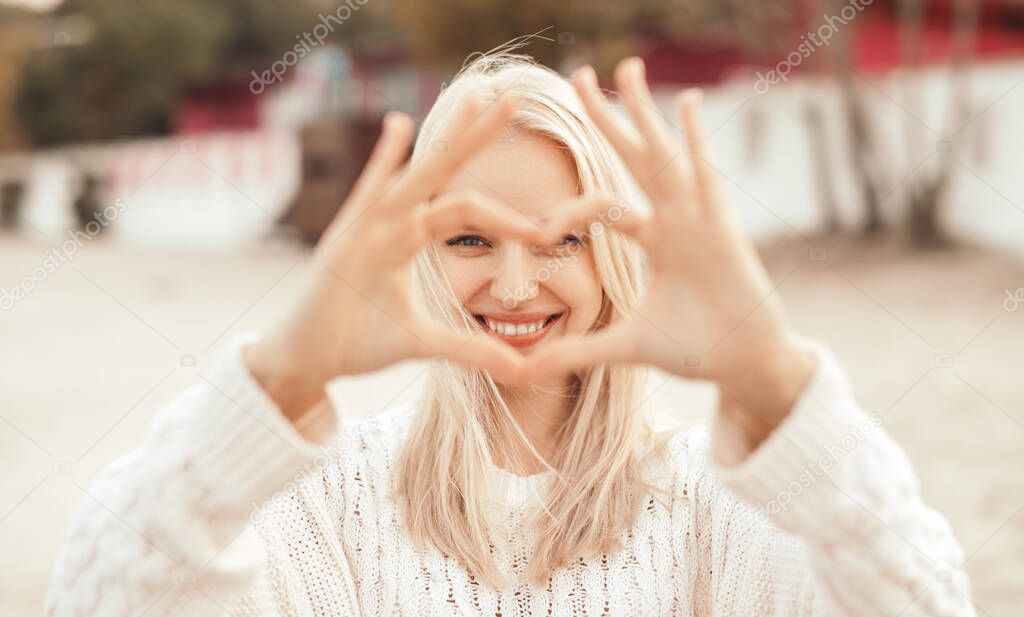 Happy romantic young woman looking at camera and showing heart gesture on street