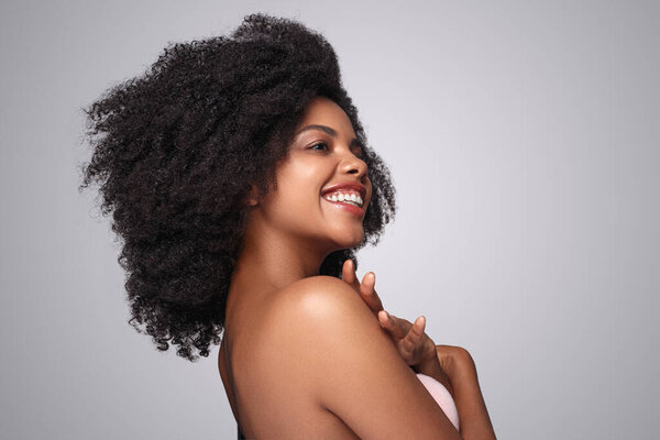 Side view of optimistic African American female with curly hair cheerfully smiling and looking away while enjoying cleanliness of skin of shoulders after spa procedure, against gray background