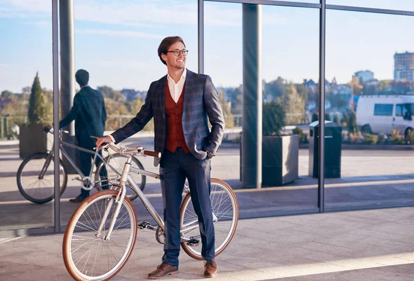 Joven hombre de negocios positivo con bicicleta de pie cerca del edificio moderno — Foto de Stock