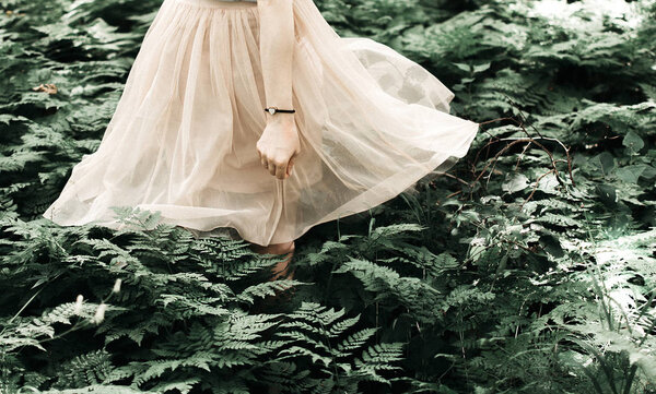 Girl in pink dress walks in the woods among ferns.