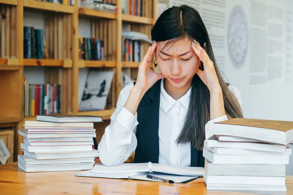 Cute Asian girl sitting in a library, surrounded by books, thinking about studying. Headache, migraine, stress after study. Teen student prepares for exams, keeps records feels bad, sick.