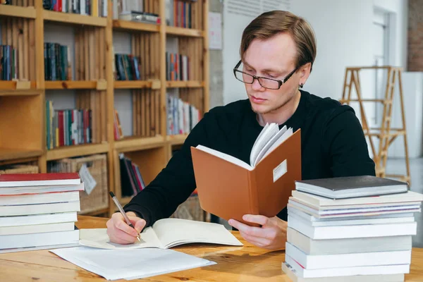 A young man in a black sweatshirt and glasses, a student or teacher, reads a book in the library, and writes information into a notebook. studies information, prepares for exams