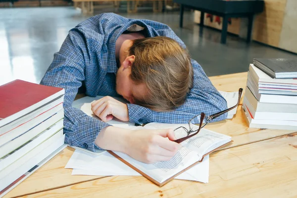 a man in a blue plaid shirt and glasses lies on the table and sleeps because of fatigue, stress, lack of sleep in a university library. Sitting at the table, preparing for exams, looking for information