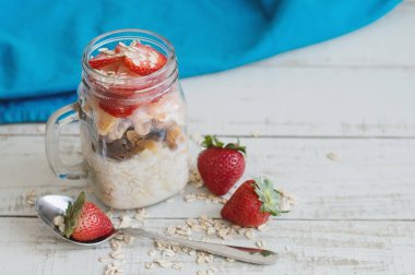 Gecede yulaf bir yüksek cam kavanoz, kaşık ve mavi mutfak havlu ile ahşap masa üzerinde bütün çilek çilek ile