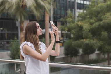 Genç Avrupalı kadın kahverengi saçlı ve beyaz rahat tarzı bluz cep telefonu ile fotoğraf çekmek. Selfie açık havada alarak kadın