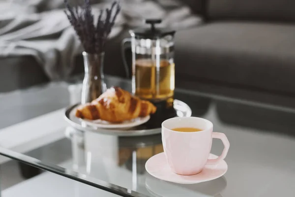 Pembe fincanda bitki çayı, taze kruvasan, çay demliği ve oturma odasındaki kahve masasında servis edilen kurutulmuş lavanta buketi. Kahvaltı konsepti