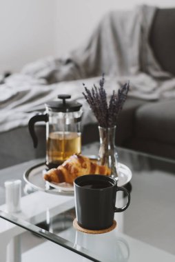 Siyah fincanda bitki çayı, kruvasan ve çay demliği metal tepside kurutulmuş lavanta buketi ile servis ediliyor. Oturma odasında kahvaltı.
