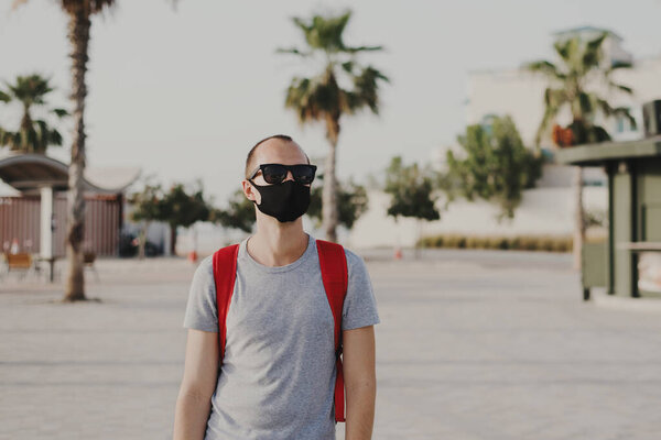 Young man in grey t-shirt, sun glasses and with backpack wearing black protective mask. Social distance and new norm concept