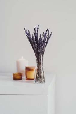 Küçük cam bir vazoda kurutulmuş lavanta ve beyaz masada aroma mumları. Basit oturma odası dekoru. Asgari düzeyde. Evde Aromaterapi