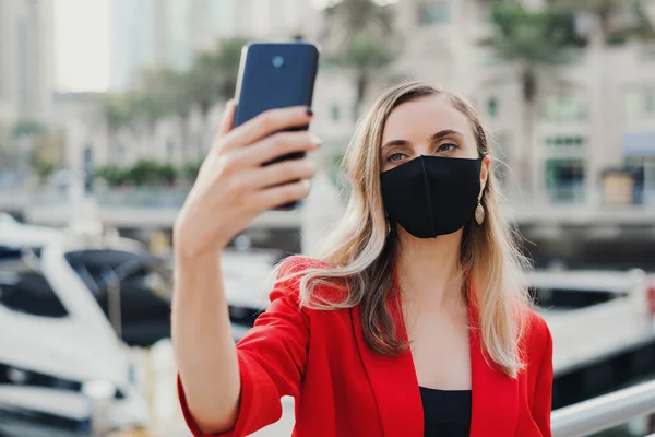 Young female model in red jacket wearing reusable face mask of black color standing in city business center and taking a selfie photo or making a video call. New normal during post Covid 19 pandemic world