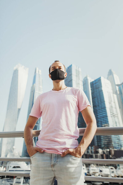 Young man in blue jeans and pink t-shirt wearing protective face mask of black color and standing in city business center with office buildings. Prevention of Covid 19