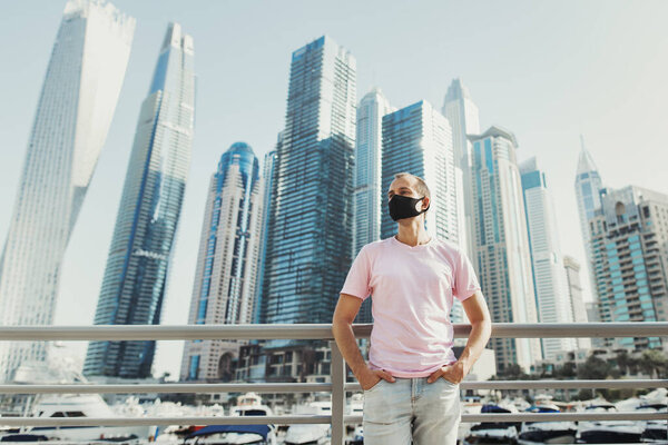 Young european man wearing pink t-shirt, jeans and protective face mask and standing near yachts and skyscrapers at city's promenade. Travel during Covid 19 or Coronavirus pandemic