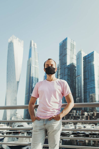 Young European man in casual outfit wearing protective face mask of black color and standing at city street with skyscrapers and office buildings. Travel during Covid 19 pandemic