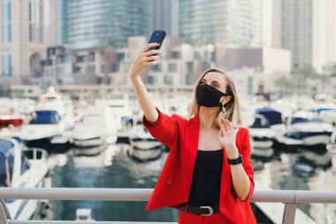 Kırmızı takım elbiseli, siyah renkli koruyucu maske takan genç bir kadın şehir merkezindeki yatların ve gökdelenlerin yakınında selfie çekiyor. Covid 19 ya da koronavirüs salgınında seyahat edin.