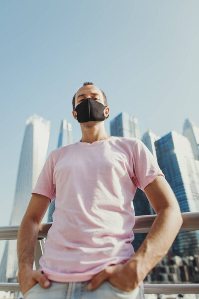 Confident young man in pink t-shirt and jeans wearing protective face mask of black color. Male tourist standing at city business center during Covid 19 pandemic