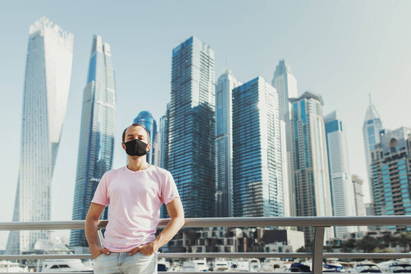 European male tourist in pink t-shirt and jeans standing at the promenade near yachts and skyscrapers and wearing protective face mask. New normal concept