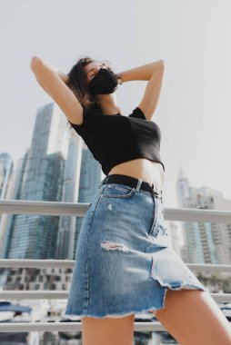 Dışarıda koruyucu yüz maskesi takan bir kadın model. Kot etekli kadın, siyah tişört ve bez solunum cihazı. Yeni normal