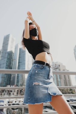 Yeniden kullanılabilir koruyucu maskeli genç bir kadın şehir caddesinde duruyor. Yeniden kullanılabilir yüz maskesi olan sıradan bir kıyafet. Hava kirliliği ve virüs koruma kavramı