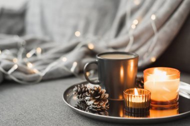 İki yanan aroma mumu, siyah kupada kahve ve yuvarlak metal tepside servis edilen dekoratif çam kozalakları. Sıcak bir atmosfer. Hygge konsepti