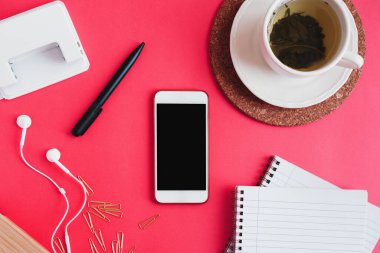 Akıllı telefon, bir fincan yeşil çay, defterler, kalem, ataç, delikli delgi ve pembe arka planda kulaklık, düz yatak. Eğitim ya da çalışma konsepti
