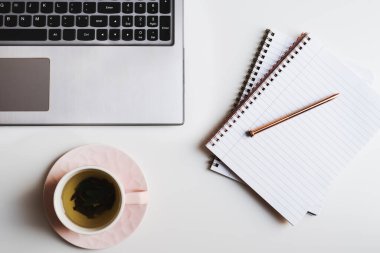 Laptop klavyesi, temiz sayfalı defterler, beyaz ofis masasındaki pembe bardakta yeşil çay, üst manzara.