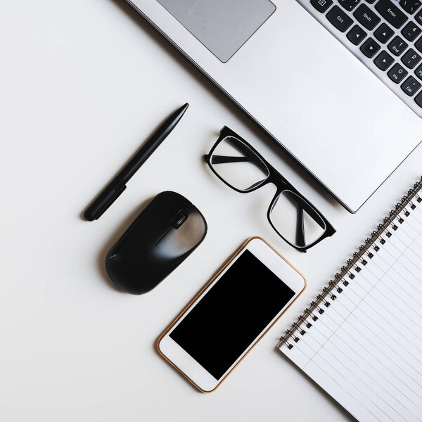 Square flat lay with laptop keyboard, notebook, reading glasses, pen, computer mouse and mobile phone on white office desk