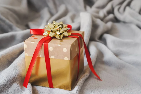 Christmas gift box of golden color with red ribbon and golden bow on gray blanket. Christmas present