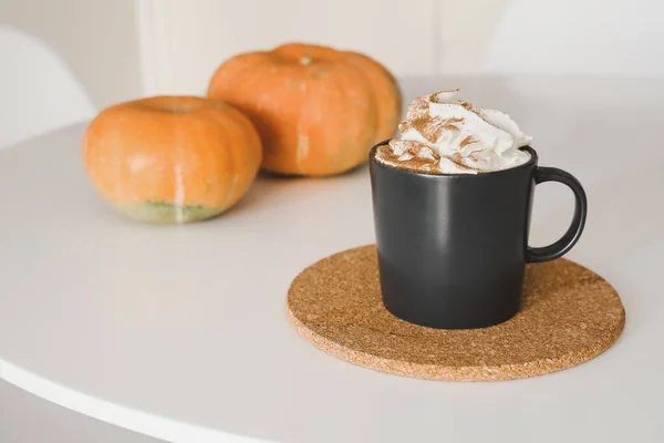 Sütlü kremalı balkabağı aromalı latte ve siyah kahve fincanında mantar altlığı ve beyaz masada iki küçük balkabağı.