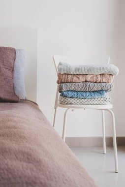 Yatak odasında duran beyaz sandalyede yün kazak yığını. Örgü kıyafetlerine dikkat et. Soğuk mevsim için sıcak kıyafetler