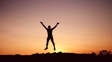 Gün batımında tepede zıplayan mutlu adamın silueti. Mutlu ve başarılı insan akşamları ellerini kaldırarak dağın tepesine atlıyor..