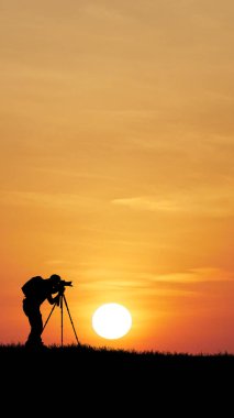 Canlı bir günbatımına karşı silueti olan bir fotoğrafçı, batan güneşin güzelliğini üç ayaklı bir kamerayla yakalıyor..