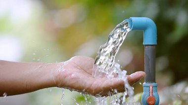 Açık mavi bir musluktan su yakalayan bir elin yakın çekimi, yemyeşil, yemyeşil bir zemin üzerine kurulmuş..