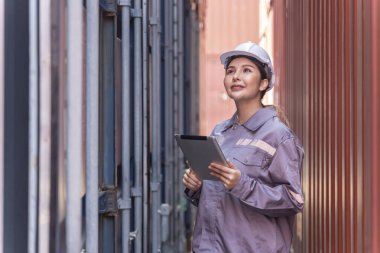 Güvenlik üniformalı genç Asyalı kadın. Tablet bilgisayar kullanıyor. Nakliye deposundaki kargo konteynırında malları kontrol ediyor.