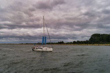 A sailboat with blue sails glides smoothly on the water, surrounded by dark clouds overhead. The shoreline is visible in the distance as the light begins to dim.