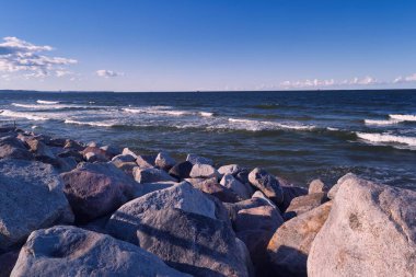 Kıyı şeridinde kıyı boyunca dağılmış büyük, pürüzsüz kayalar bulunur. Dalgalar parlak mavi bir gökyüzünün altında yavaşça süzülüyor dağınık bulutlarla, sakin bir sahil atmosferinin özünü yakalıyor..