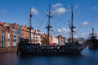 A black pirate ship is docked along the waterfront in Gdansk. The vibrant buildings lining the canal reflect the city's rich maritime history under a clear blue sky.