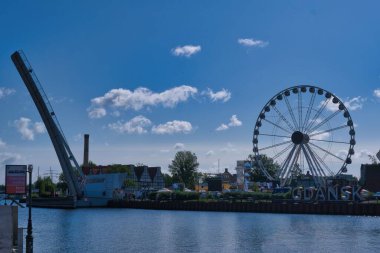 The vibrant waterfront of Gdansk showcases a large ferris wheel along with a drawbridge. Green trees and colorful buildings surround the serene water under a bright blue sky.