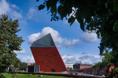 A striking angular red building stands prominently against a backdrop of fluffy clouds and blue sky.