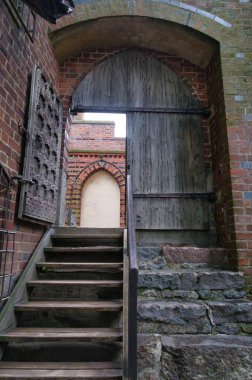 Taş basamaklar, aşınmış tuğla duvarlarla çevrili kemerli ahşap bir kapıya doğru yükseliyor. Tarihi mimaride rustik ayrıntılar ve eski dokular sergileniyor.