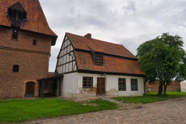 A charming half timbered house with a red tiled roof is located next to a tall brick tower. The street is cobbled and lined with greenery under an overcast sky.